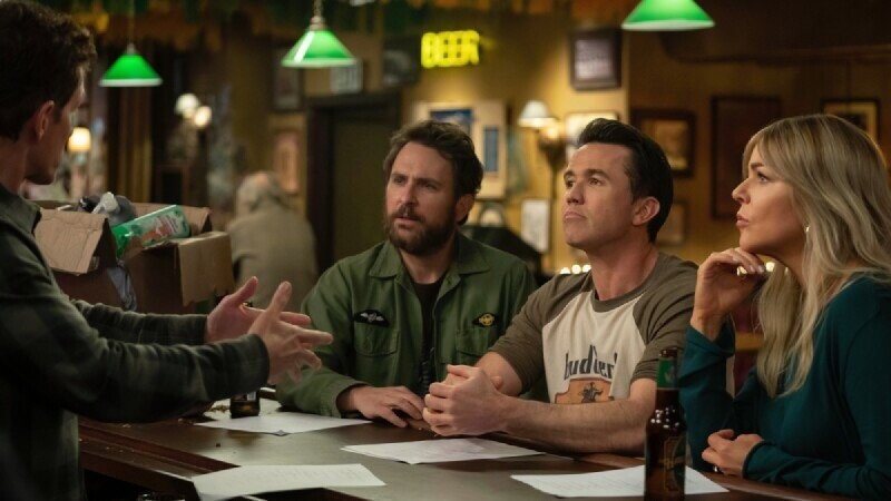 Four adults are seated at a bar counter in a dimly lit pub, engaged in an intense conversation. Three of them-a man wearing a green jacket, a man in a beige and brown t-shirt, and a woman with long blonde hair in a teal top-are looking at the fourth person, whose hands are gesturing animatedly as he speaks. Papers and beer bottles are spread out on the bar, with warm lighting and green hanging lamps creating a cozy atmosphere.