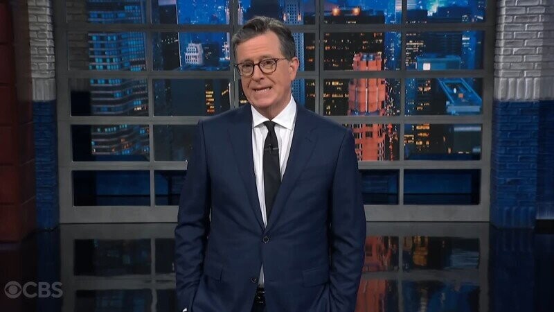 Man wearing a dark blue suit indoors in front of large window with a night cityscape