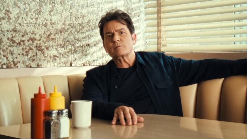 A man with dark hair is sitting alone in a booth at a diner, leaning back with one arm resting along the top of the seat. He is wearing a dark shirt and jacket, and his expression is serious or contemplative. On the table in front of him are a white coffee mug, a salt shaker, a pepper shaker, and bottles of ketchup and mustard. The booth is upholstered in a light beige color, and behind him there is a window with horizontal blinds partially open, letting in soft light. The wall next to the window has a patterned design.