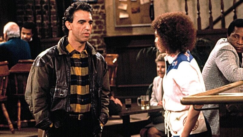 The image shows a man and a woman engaged in conversation inside a dimly lit bar or pub with brick walls and wooden furniture. The man is wearing a brown leather jacket over a yellow and black checkered shirt, while the woman has curly hair and is dressed in a white shirt with blue trim and an apron, suggesting she might be a waitress. In the background, other patrons are seated and chatting near a wooden staircase and bulletin board.